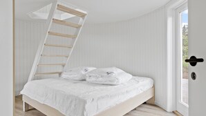 Ein Schlafzimmer mit schrägverdeckter Decke, einem Bett mit weißer Bettwäsche und Blick durch eine Tür.