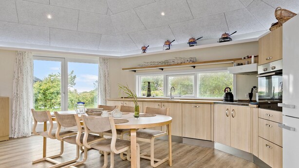 Eine moderne Küche mit Holzschränken, eine Essecke mit rundem Tisch und Stühlen sowie große Fenster mit Blick auf Grünflächen.