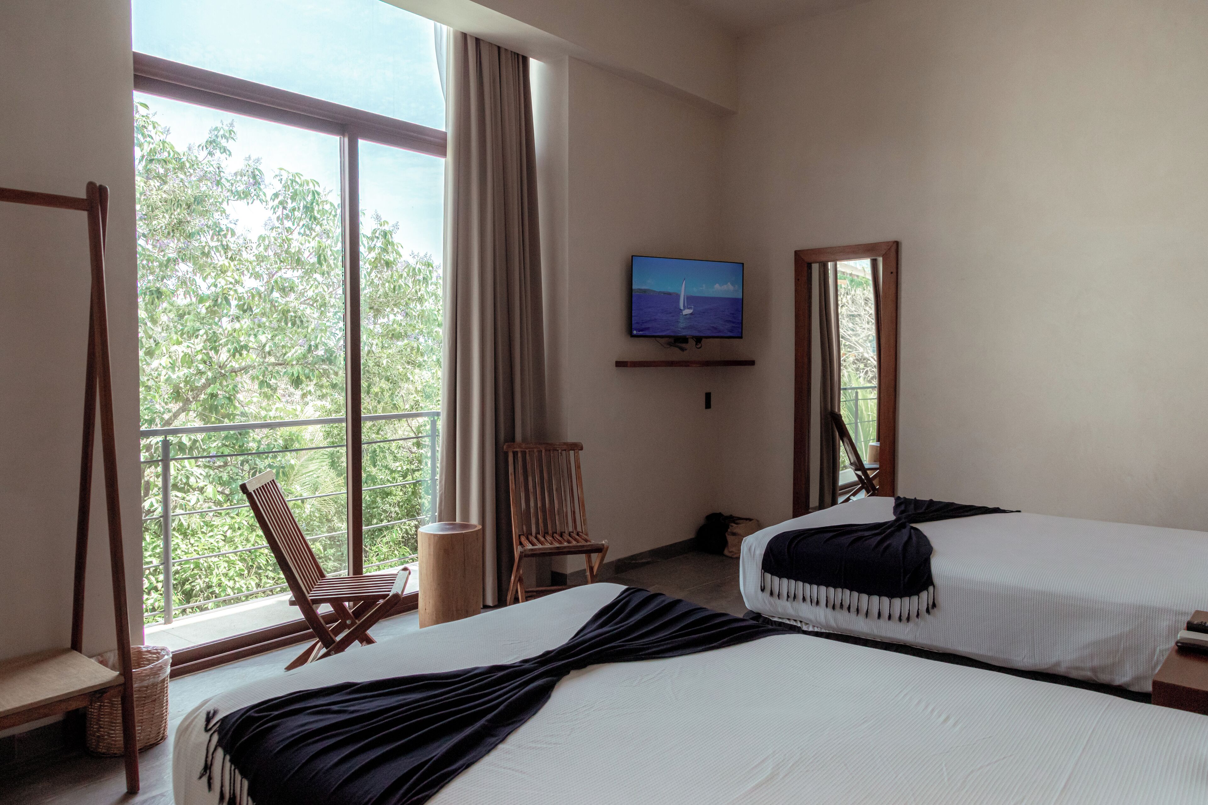 Ein Hotelzimmer mit zwei Betten, einem an der Wand befestigten Fernseher, einem Balkon mit Stuhl und Blick auf Bäume.