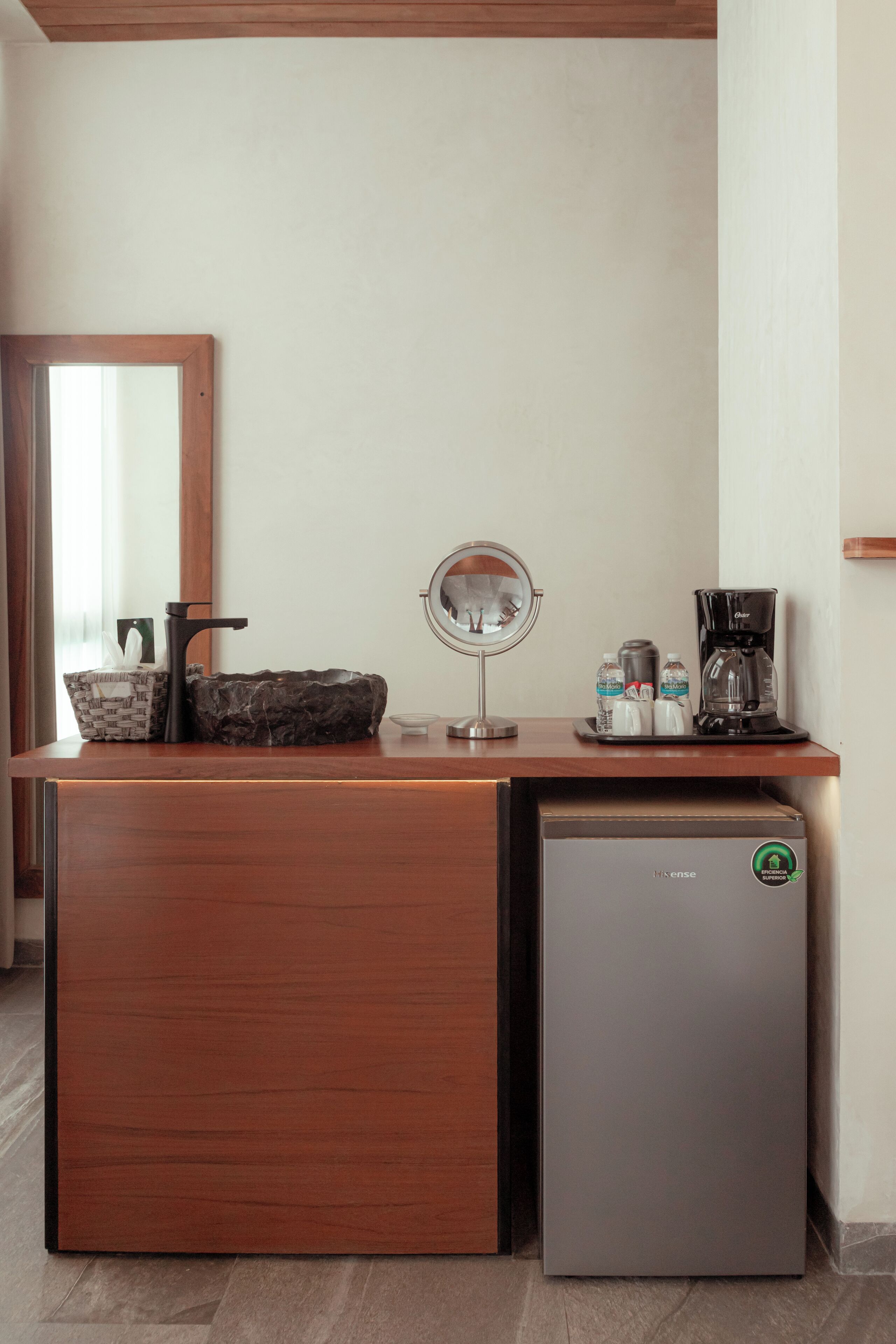 A compact kitchenette with a sink, mirror, coffee maker, and a small refrigerator.
