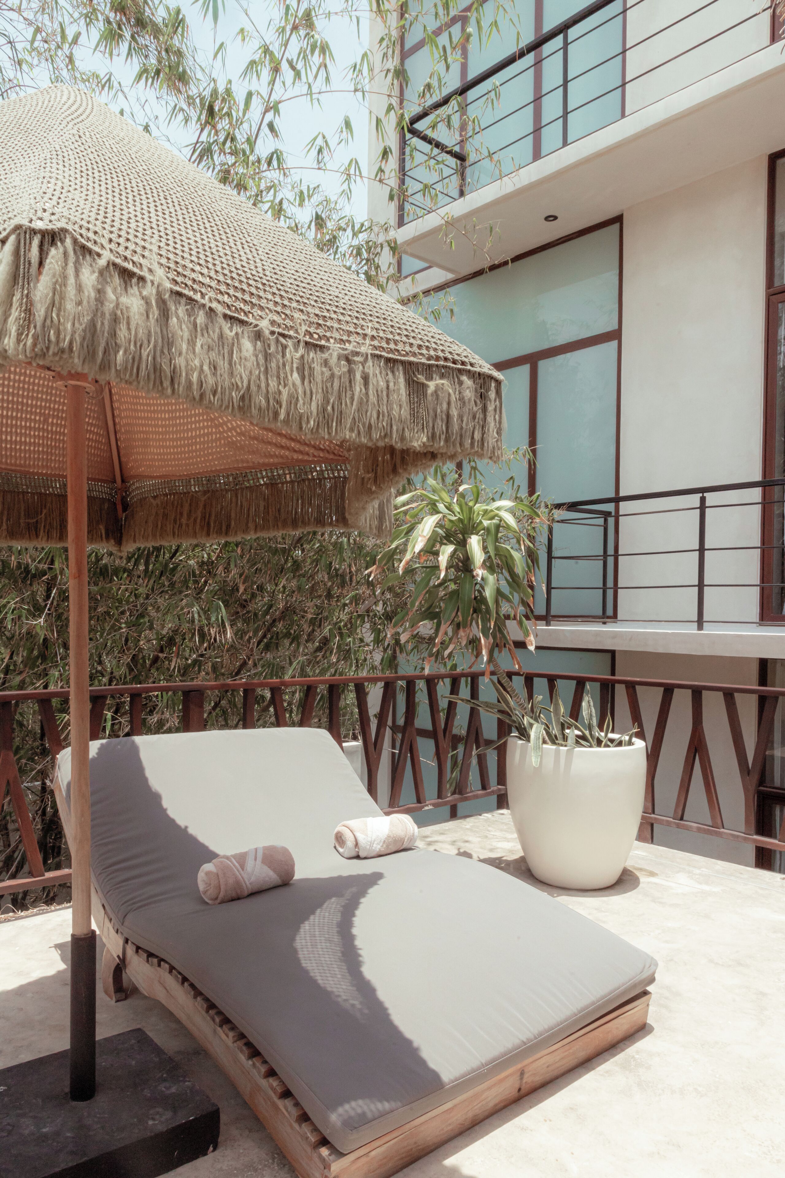 Un transat avec un parasol en osier, un pot de fleurs et une vue sur un bâtiment moderne doté de grandes fenêtres.