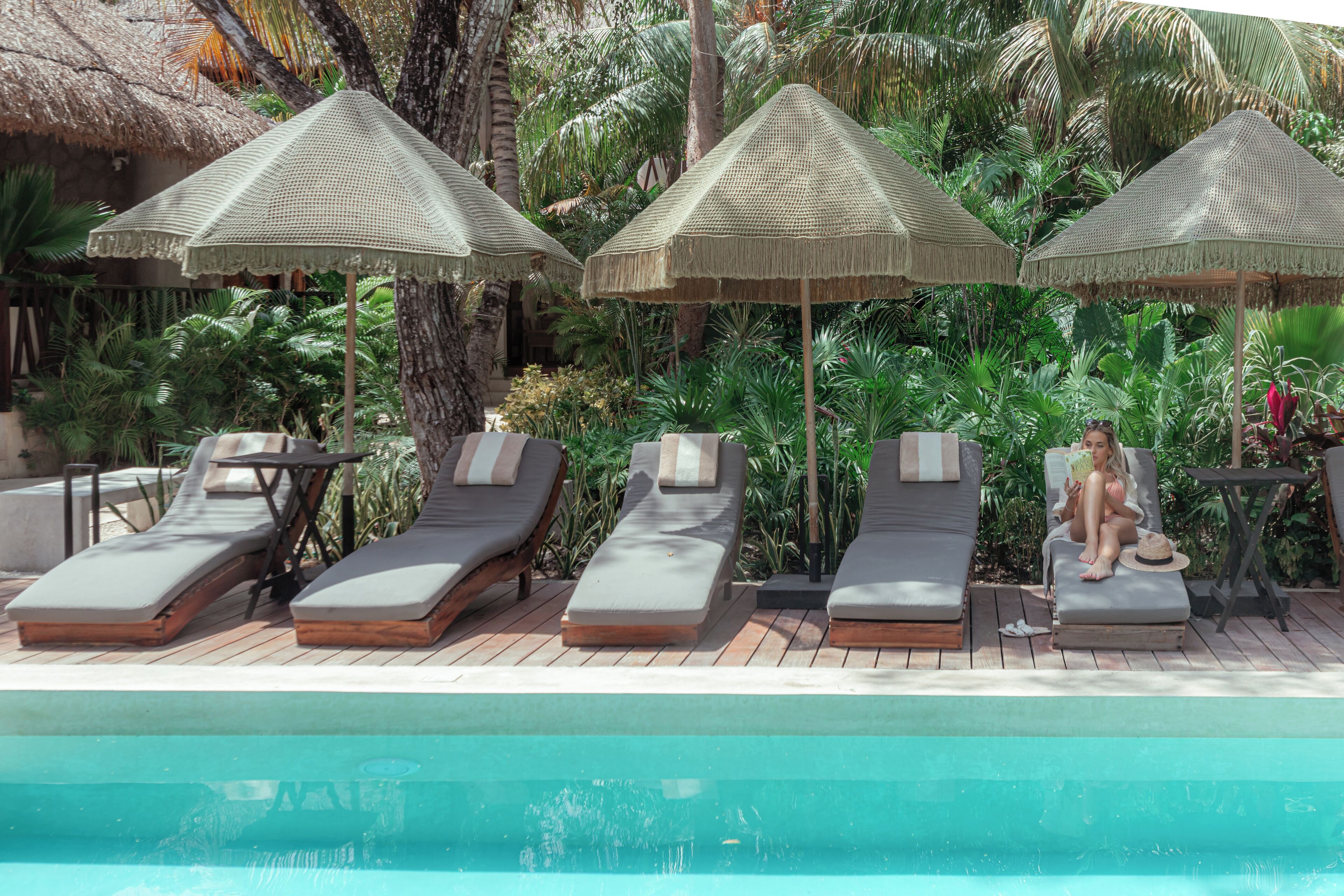A poolside area with lounge chairs and thatched umbrellas, surrounded by lush tropical plants.