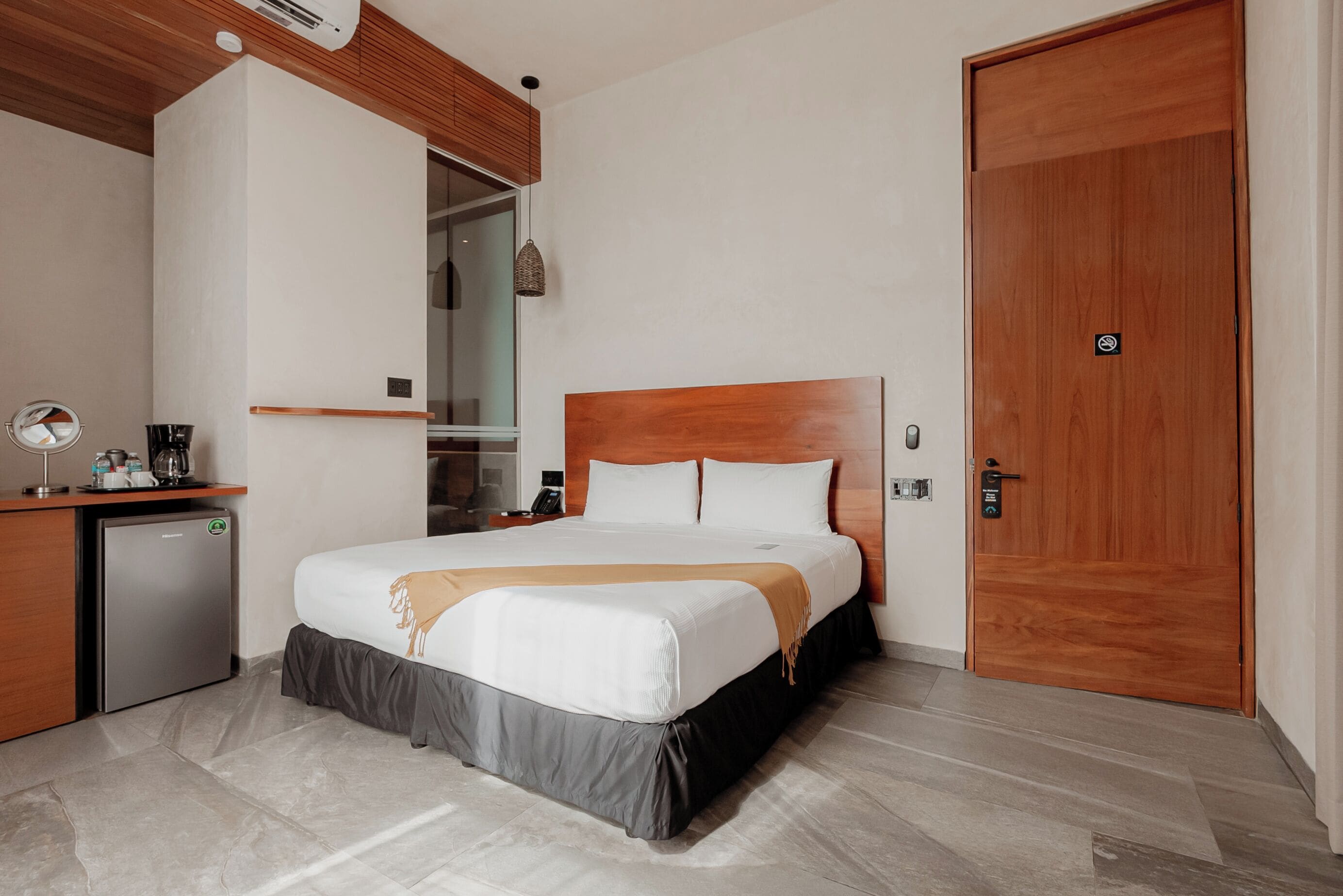 A hotel room with a bed, a wooden headboard, a wardrobe, a desk with a coffee maker, and a small refrigerator.