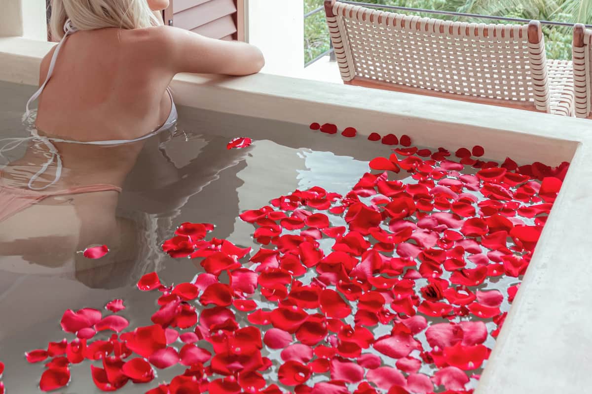 A woman in a bathtub filled with water and red rose petals, with a wicker chair and a view of greenery outside.
