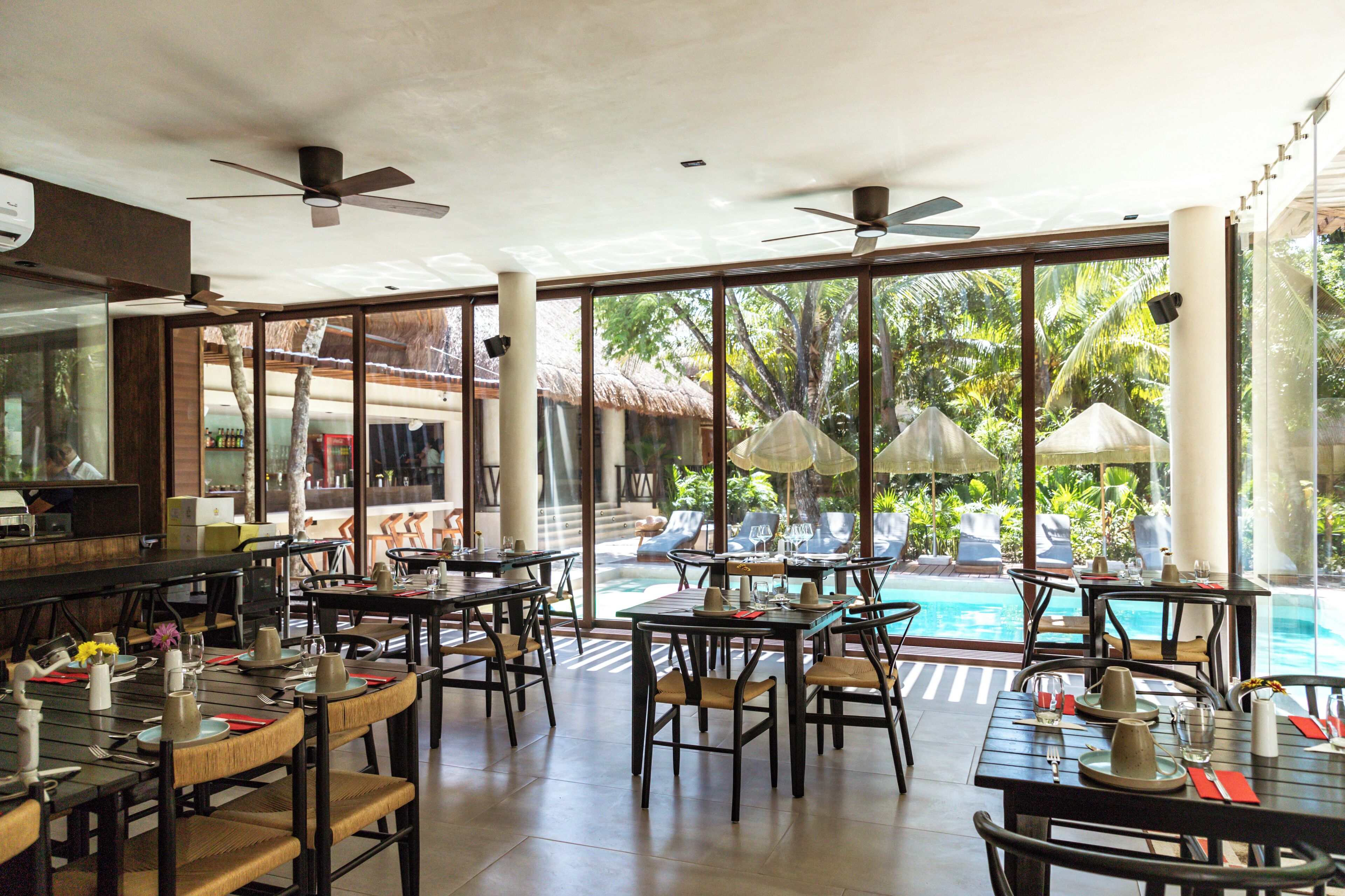 Un restaurant doté d’une terrasse avec vue sur une piscine, de grandes fenêtres et de ventilateurs de plafond.