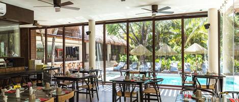 Un restaurant doté d’une terrasse avec vue sur une piscine, de grandes fenêtres et de ventilateurs de plafond.