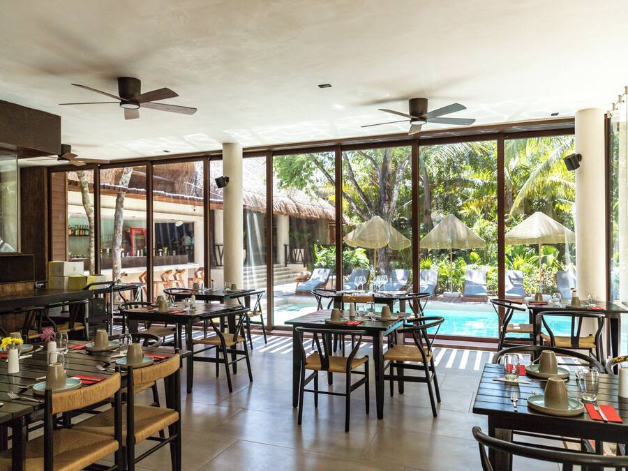 Un restaurante con terraza al aire libre, vista a una piscina, grandes ventanales y ventiladores de techo.