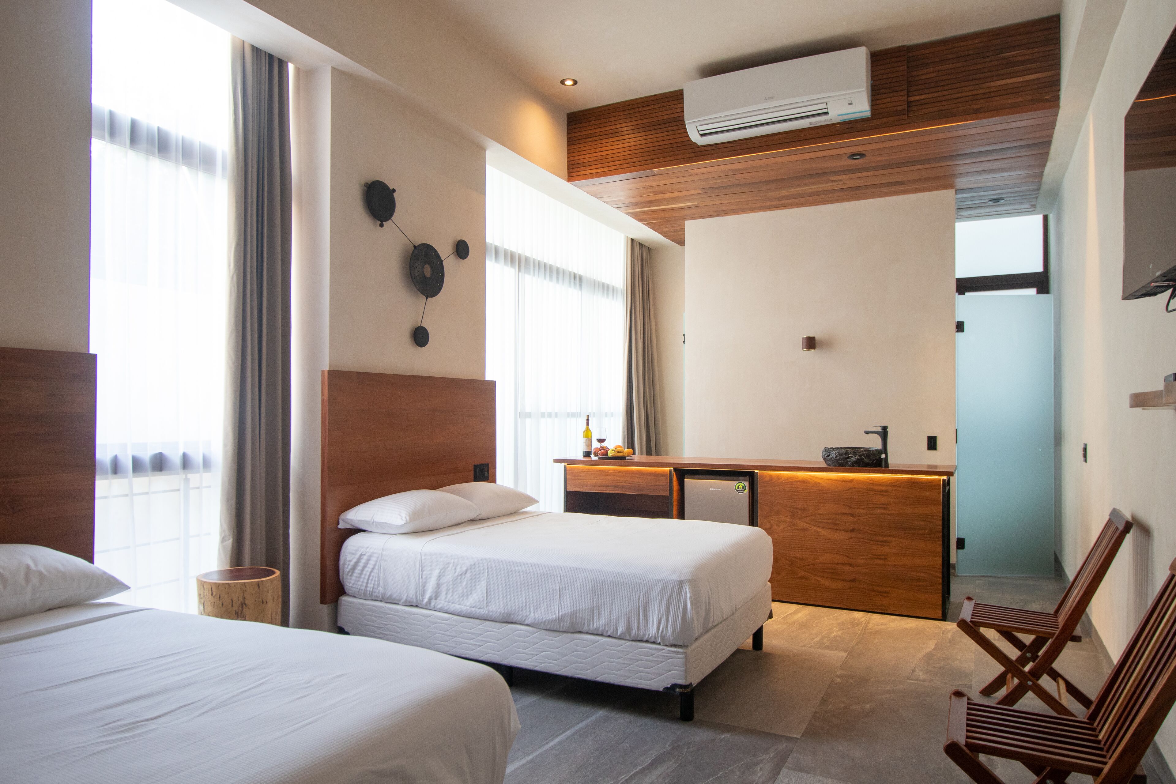 Une chambre d’hôtel avec deux lits, un bureau en bois, un petit réfrigérateur et une grande fenêtre avec des rideaux.