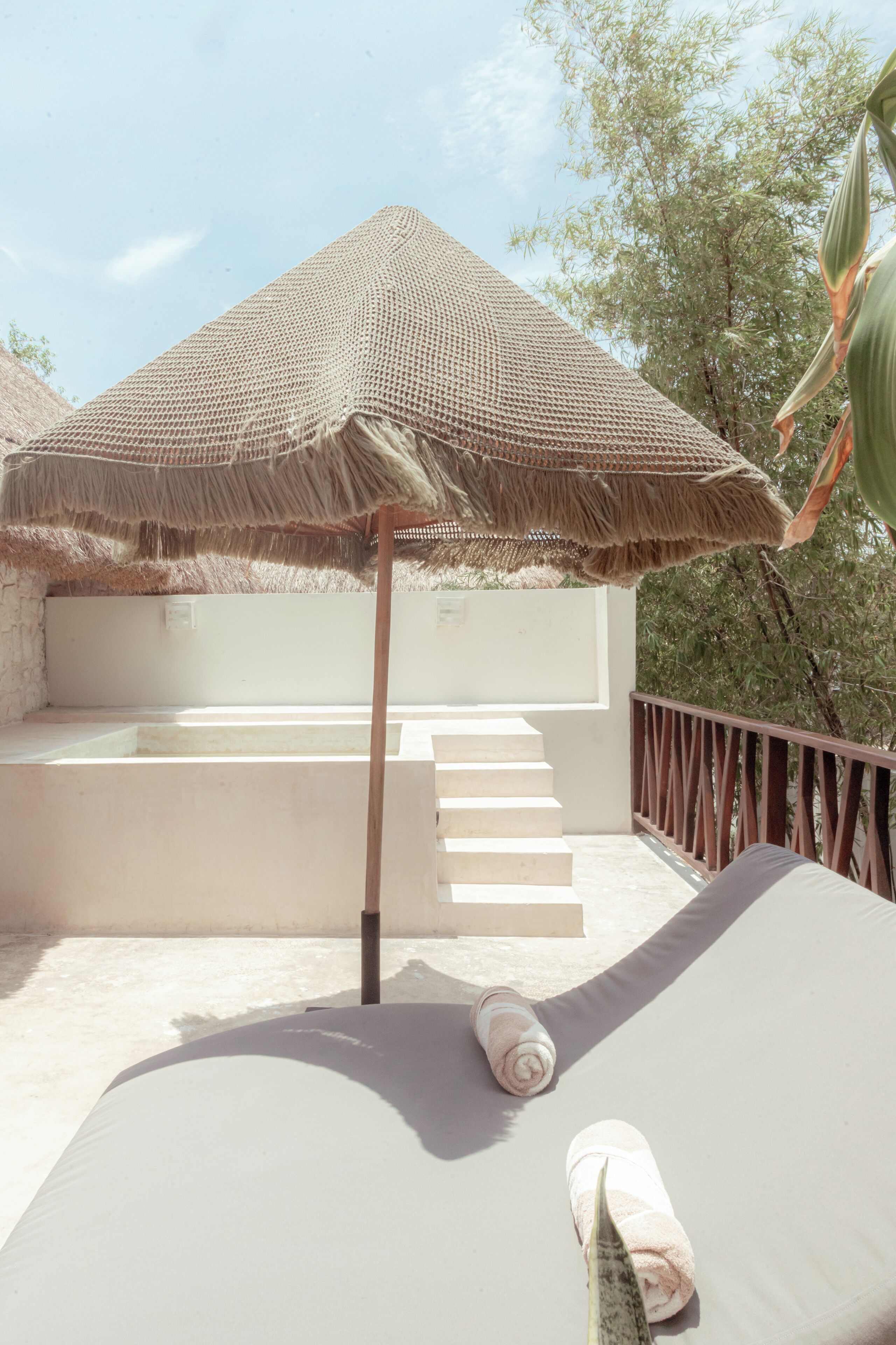 A sunlit outdoor lounge area with a thatched umbrella, a white lounge chair, and a set of stairs leading to a covered seating area.