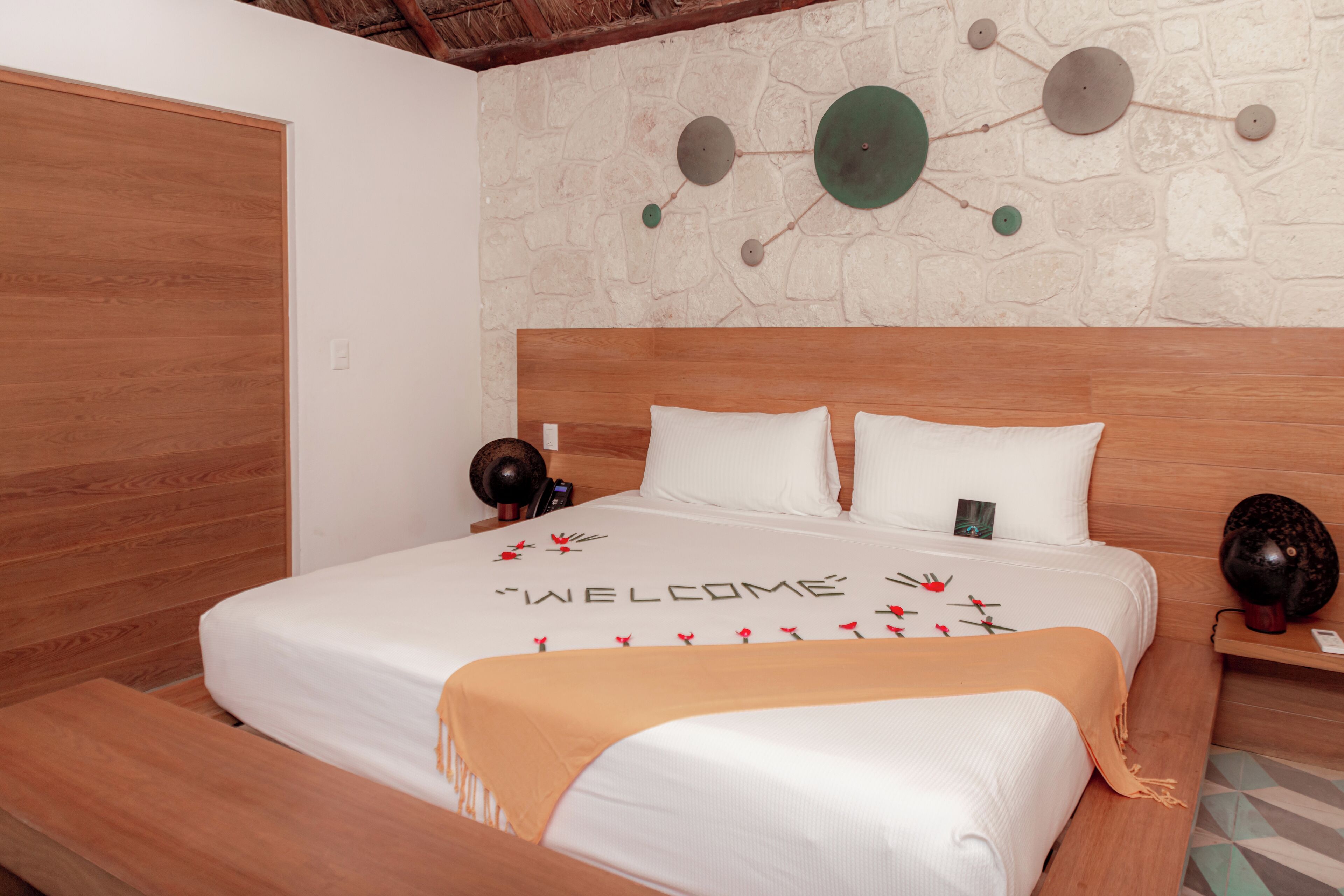 A neatly made bed with a 'WELCOME' sign, a wooden headboard, and a stone wall with decorative plates.