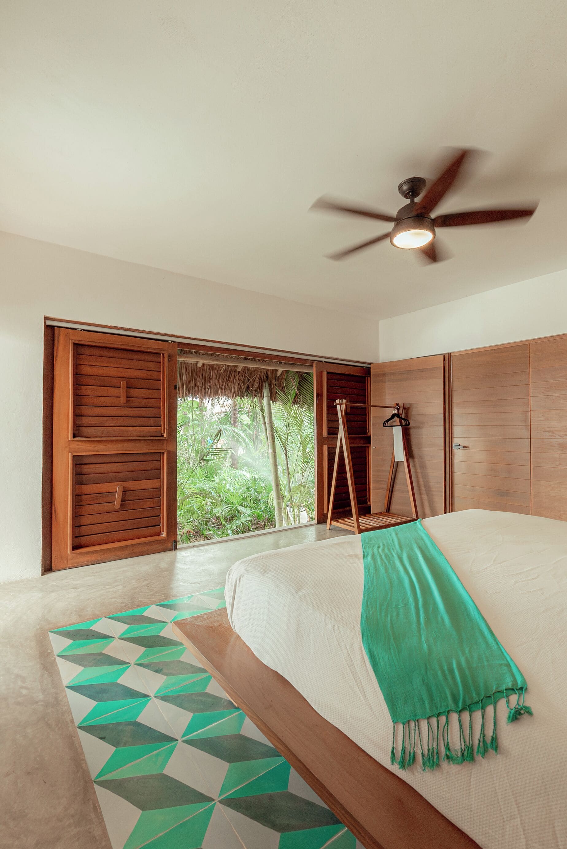A bedroom with a bed, a ceiling fan, a wooden wardrobe, and a view of greenery outside.