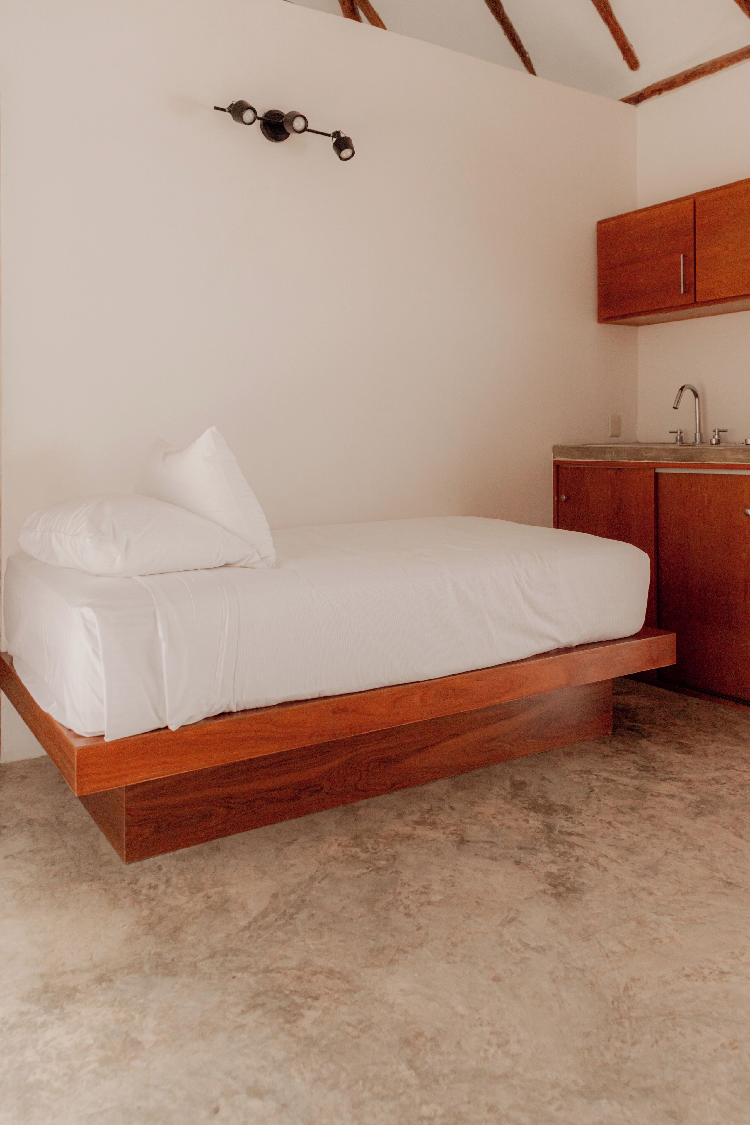 A bed with a wooden frame and white bedding, a wooden cabinet with a sink, and a wall-mounted light fixture.