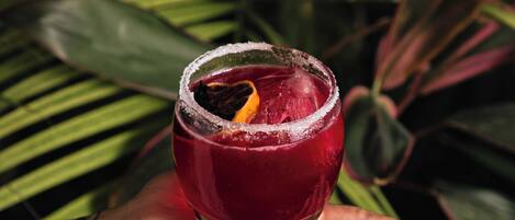 A hand holding a glass with a red drink, surrounded by green foliage.