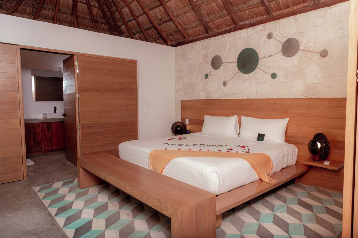 A bedroom with a wooden bed, a bench, a small bedside table, a wall-mounted clock, and a stone wall with circular decorations.