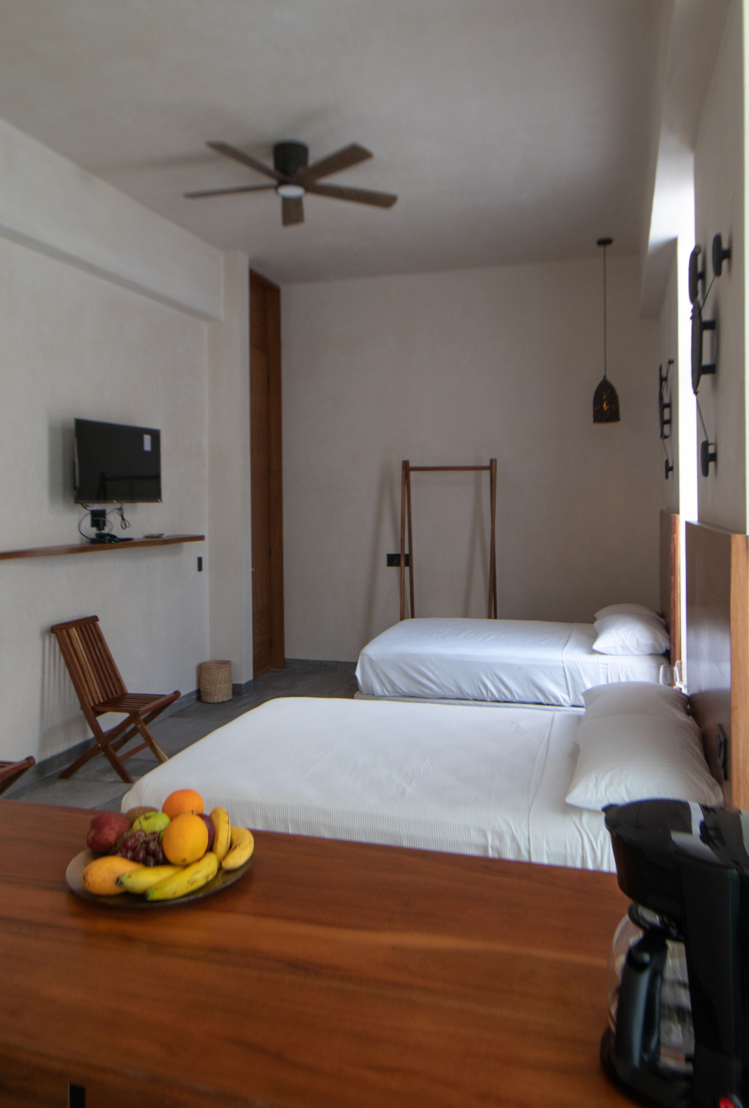 A hotel room with two beds, a wooden desk with fruit, a chair, a television, and a ceiling fan.