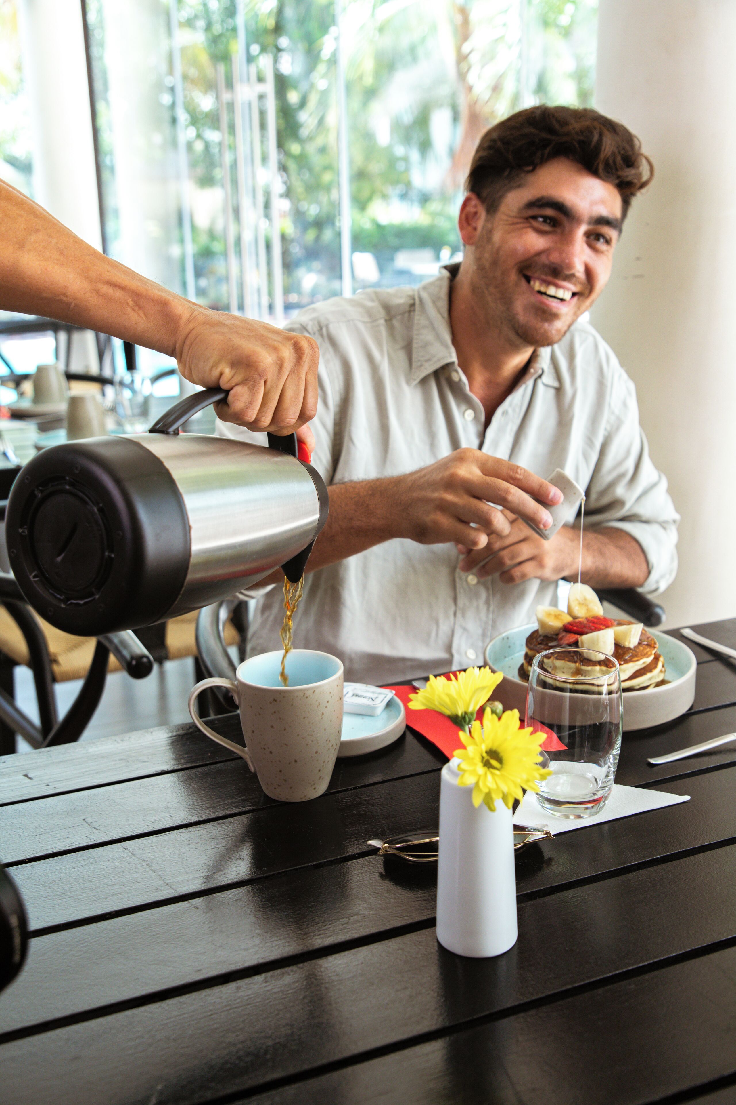 A person pouring a beverage into a cup on a table with a flower vase, a plate with food, and a glass of water.