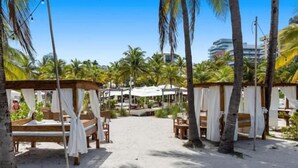 A beachside resort with wooden beds and white canopies, surrounded by palm trees and a clear sky. - BeachFront On The Sand Unit #227 South of 5th, 335 Ocean Dr, Miami Beach (Miami Beach)