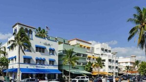 A multi-story hotel with a white facade and blue accents, featuring balconies and palm trees.