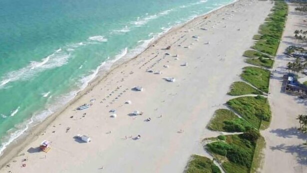 An aerial view of a beach with rows of umbrellas and sun loungers, a sandy shore, and a clear blue sea.
