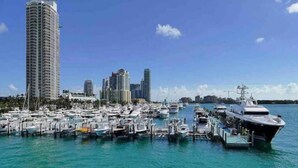 Un puerto deportivo con numerosas embarcaciones amarradas, un edificio alto a la izquierda y el horizonte de una ciudad al fondo.