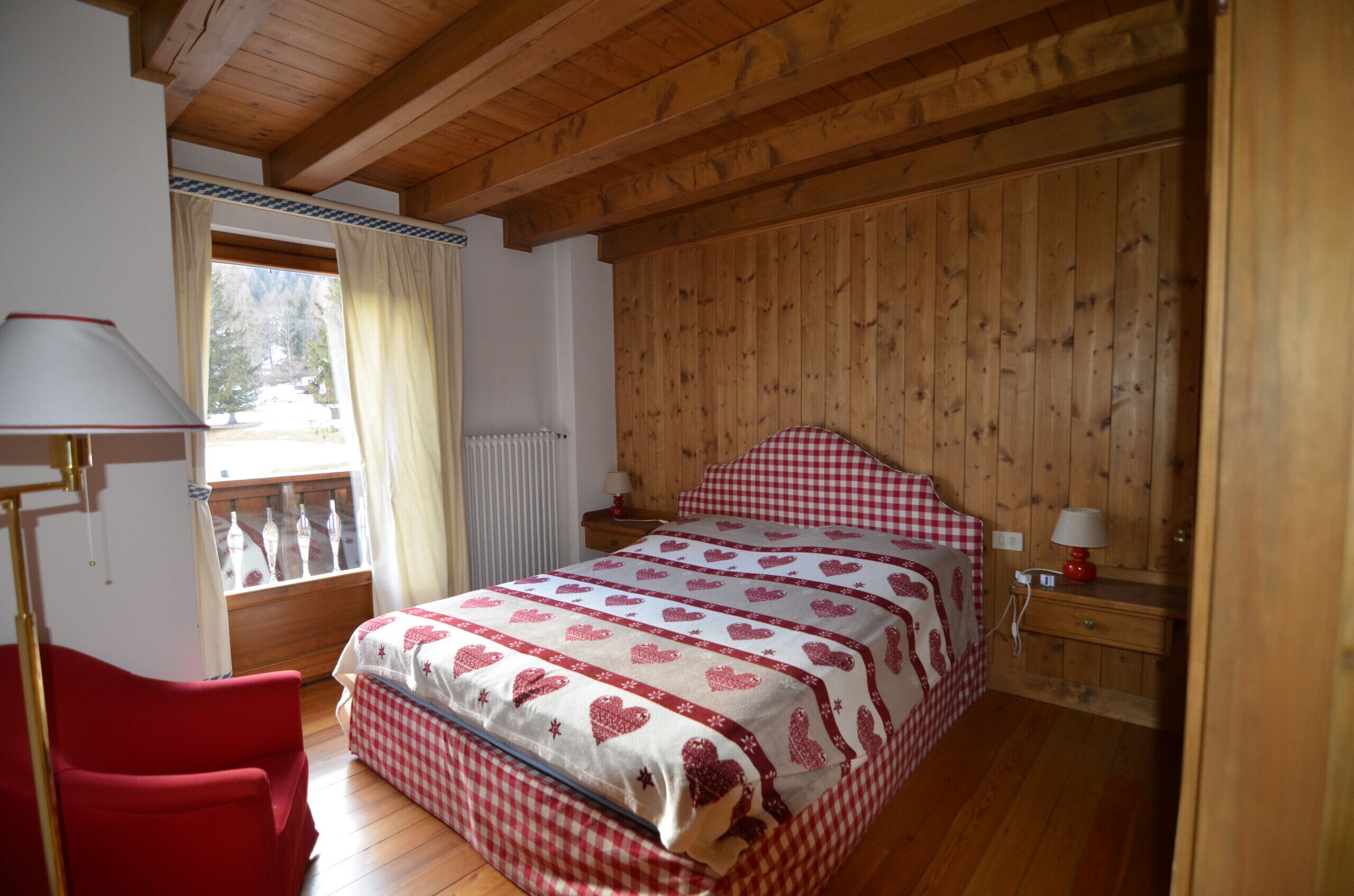 Een slaapkamer met een houten plafond, een bed met een geruit beddegoed, een rode stoel, een raam met gordijnen, en een nachtkastje met een lamp.