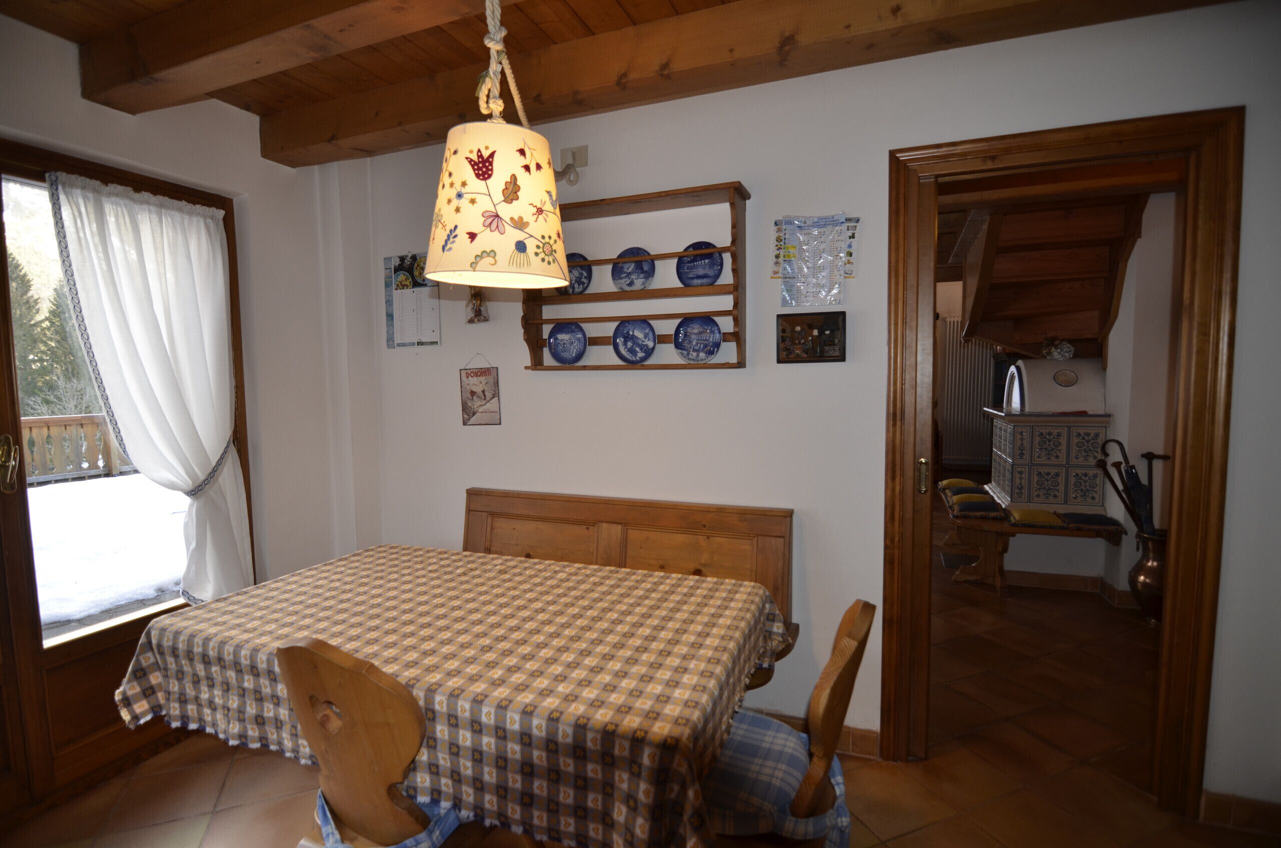 A bedroom with a wooden bed, a window with curtains, a shelf with blue plates, and a wooden ceiling.