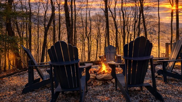 Dos sillas Adirondack mirando un fuego de campamento en un bosque al atardecer.