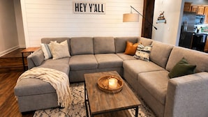Una sala de estar moderna con un sofá seccional, una mesa de centro de madera con una vela, y un cartel en la pared que dice 'HEY Y'ALL'.