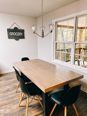 Un coin repas avec une table en bois et des chaises noires, un lustre et une fenêtre donnant sur une terrasse.
