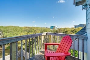 A wooden deck with a red Adirondack chair, overlooking a lush green landscape and a distant house.