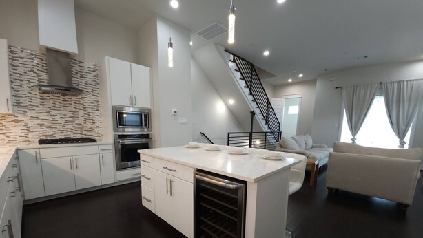 A modern kitchen with white cabinetry, a central island, and stainless steel appliances. There is a staircase leading to a living area with a sofa and curtains. - "modern 3bed Townhome. Near Nrg" (Houston)