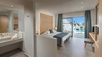 A modern hotel room with a large bed, a bathroom with a sink and mirror, a desk with a TV, and a balcony with a pool and lounge chairs.