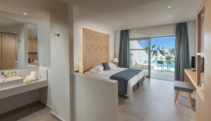 A modern hotel room with a large bed, a bathroom with a sink and mirror, a desk with a TV, and a balcony with a pool and lounge chairs. - Blue Lagoon Ocean (Kos)