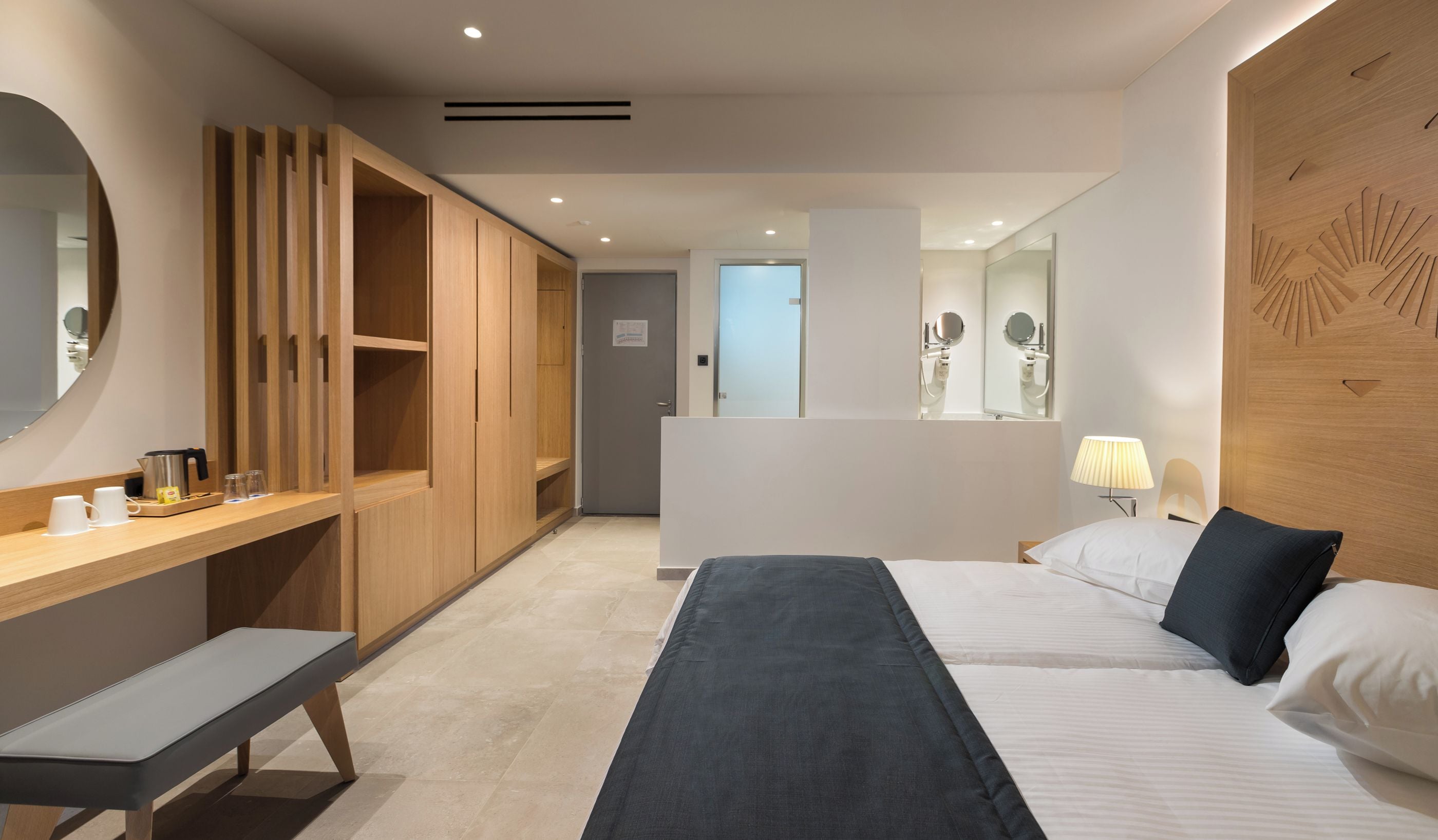 A modern hotel room with a wooden wardrobe, a desk with a kettle, a bed with white and dark pillows, and a bedside lamp.