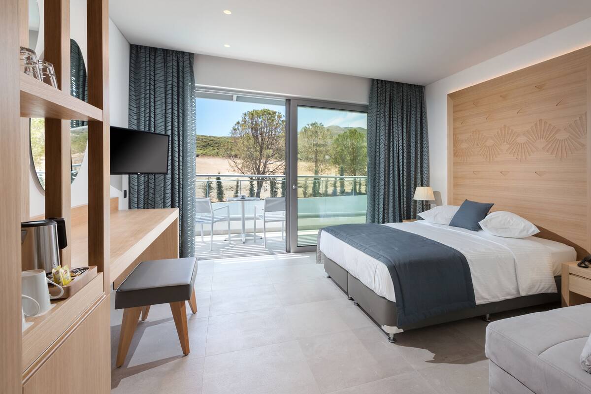 A modern hotel room with a large bed, a desk, a chair, and a balcony with a view of the outdoors.