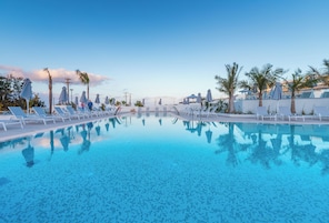 A swimming pool with lounge chairs and umbrellas, surrounded by palm trees and a clear sky.