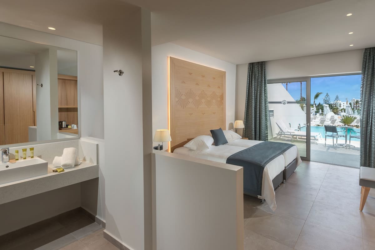 A modern hotel room with a large bed, a bathroom with a sink and mirror, a desk with a TV, and a balcony with a pool and lounge chairs.