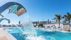 Une piscine agrémentée d’une fontaine, un ciel bleu dégagé et des chaises longues en arrière-plan.