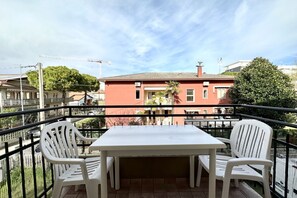 Ein Balkon mit einem weißen Tisch und zwei weißen Stühlen, mit Blick auf ein rotes Gebäude mit einem Ziegeldach und einem Schornstein.