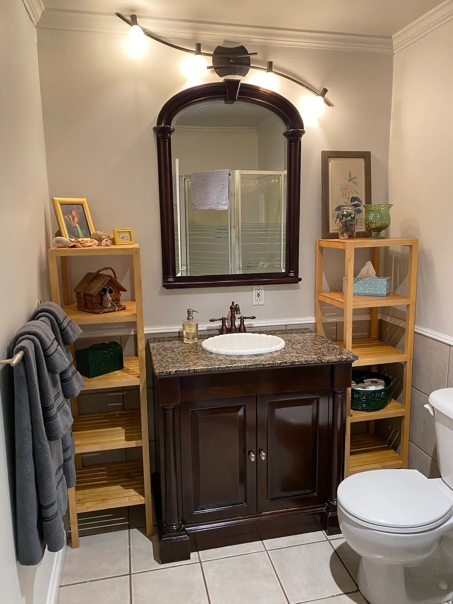 A bathroom with a dark wood vanity, a white sink, a large mirror, and a toilet. There are shelves with various items and a towel rack.