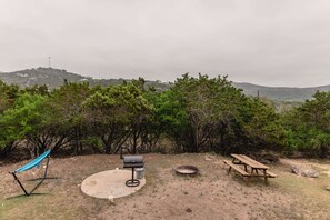 Escape to a serene oasis with a picnic table, barbecue grill, and hammock amidst lush greenery and distant hills.
