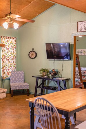 A cozy room with a wooden ceiling, a television mounted on the wall, a dining table with chairs, and a window with patterned curtains.
