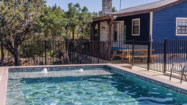 Une piscine bordée de pierres, avec une terrasse en bois et une clôture en métal.