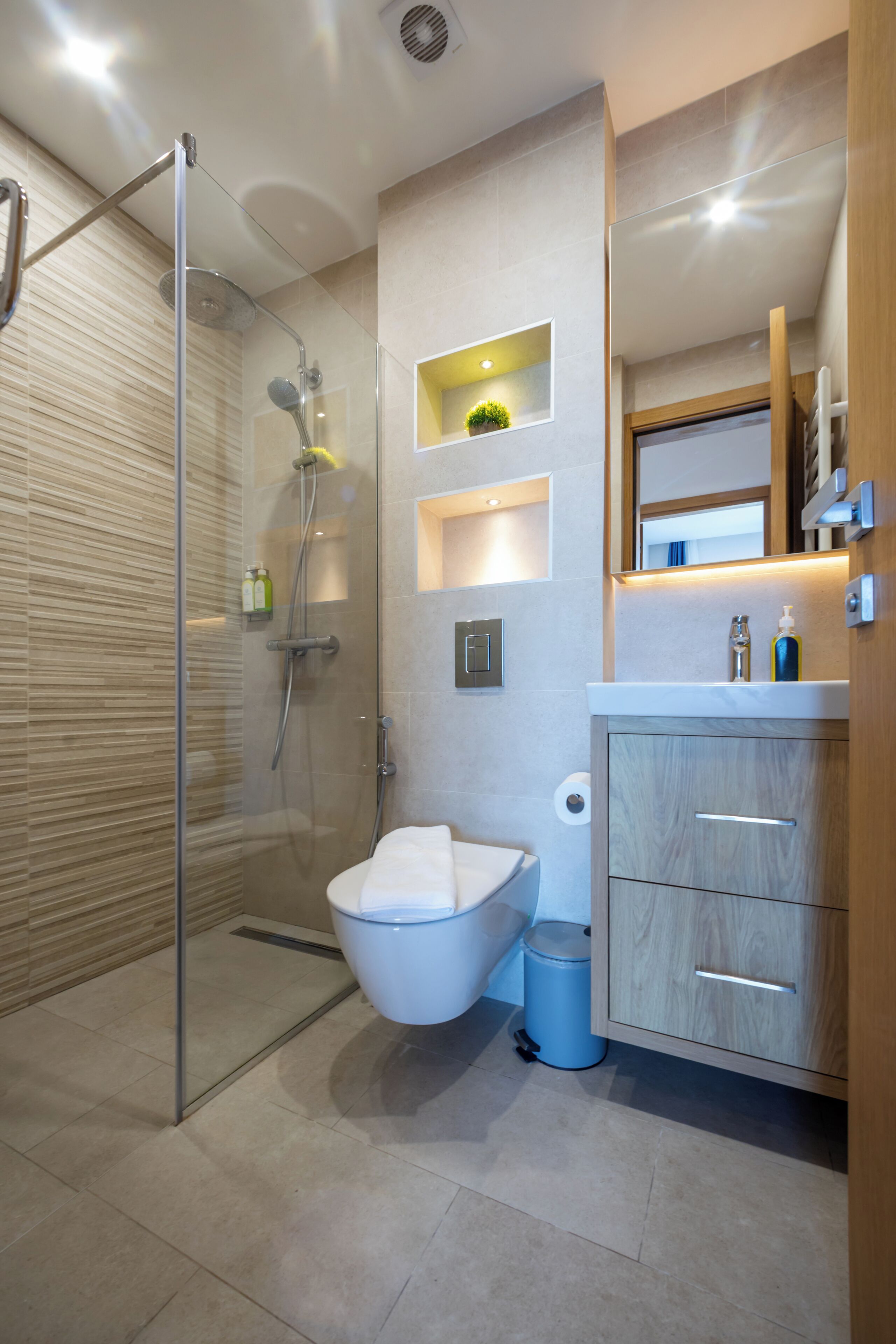 Un baño moderno con ducha de vidrio, inodoro, lavamanos con mueble de madera y una pequeña repisa con artículos de tocador.