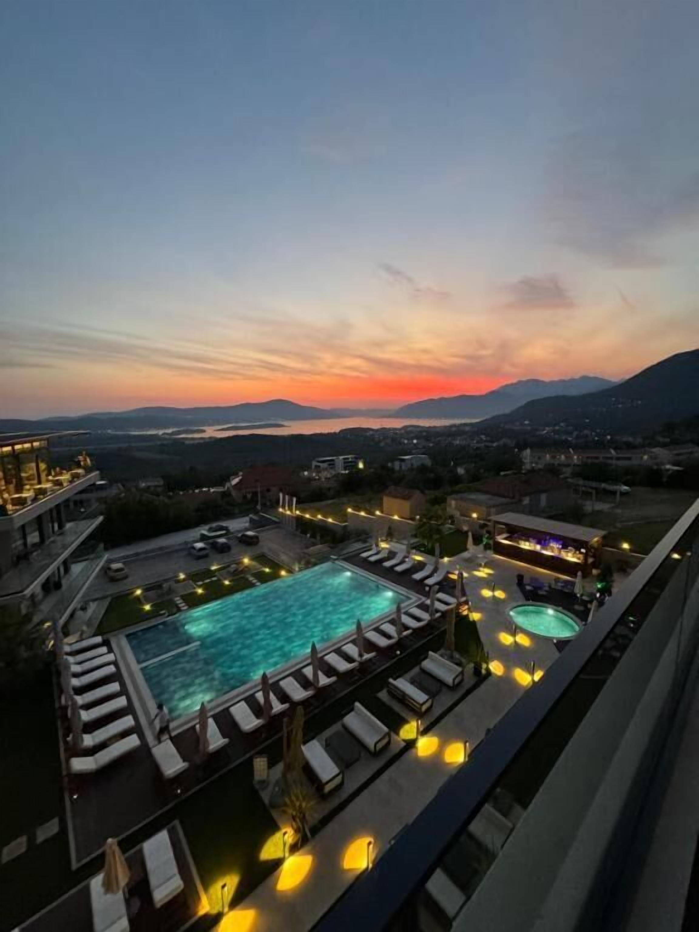 Area piscina sul tetto con diverse piscine e poltrone prendisole, circondata da edifici e vista sul tramonto.