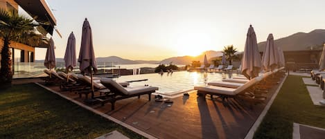 Zona junto a la piscina con tumbonas y sombrillas, una terraza de madera y vistas a la puesta de sol sobre las montañas.