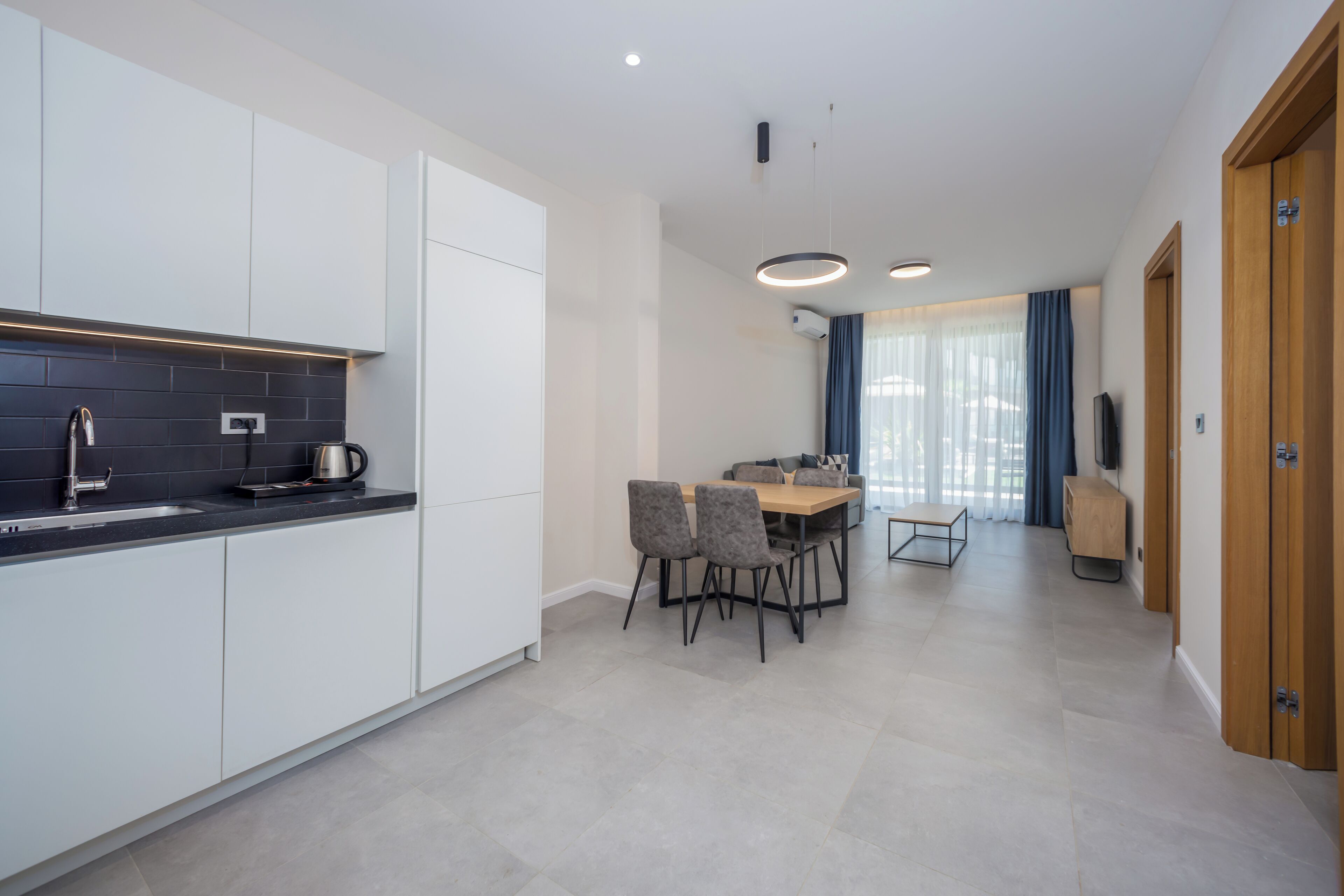 A modern kitchen with white cabinets, a black countertop, and a coffee maker. There is a dining area with a table and chairs, and a TV mounted on the wall.