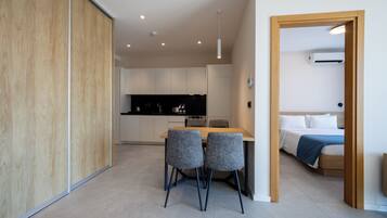 Una habitación de hotel moderna con suelo de madera, una zona de comedor con dos sillas, una cama con manta azul y una kitchenette al fondo.