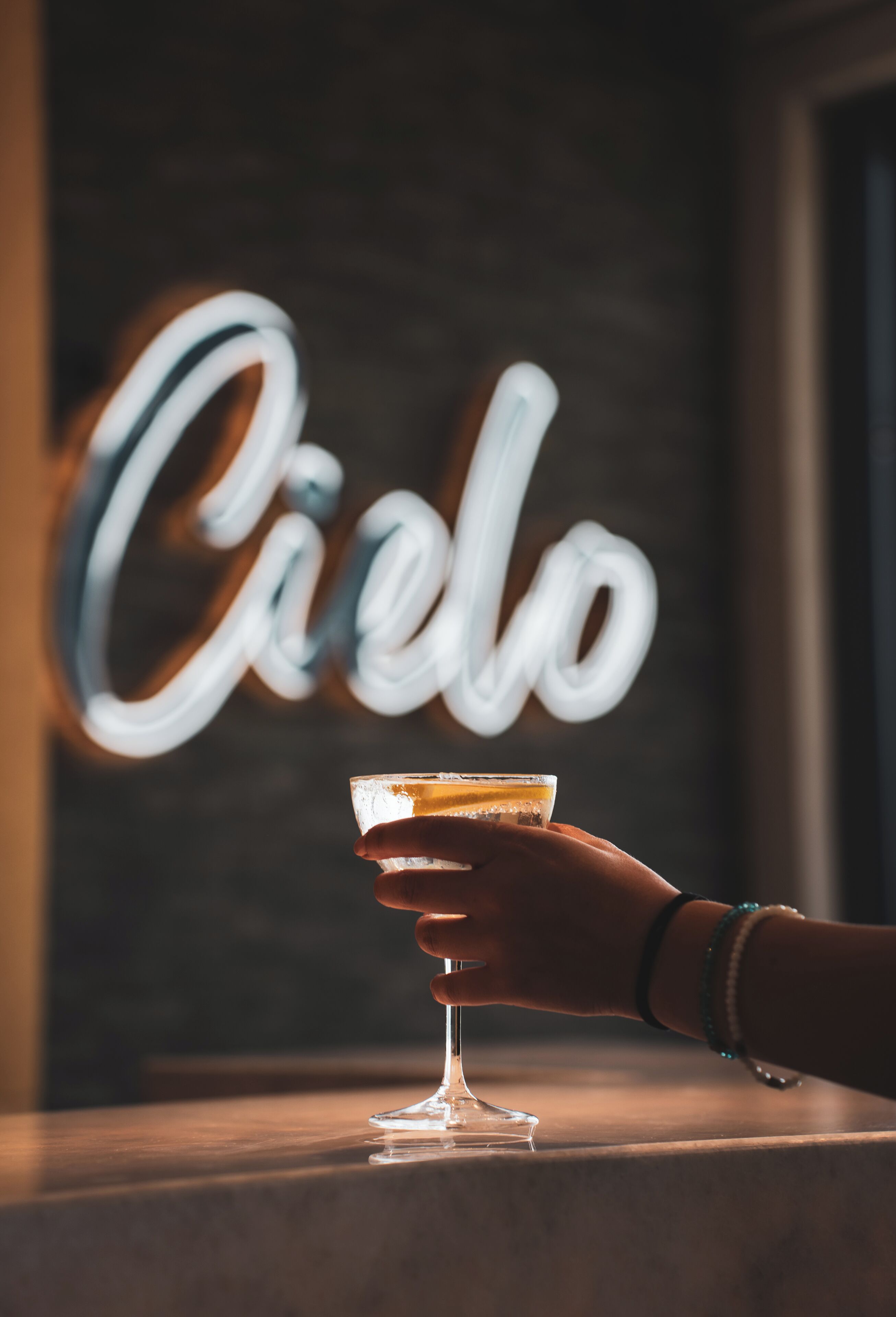 A hand holding a cocktail glass with a drink, in front of a neon sign that reads "Cielo".