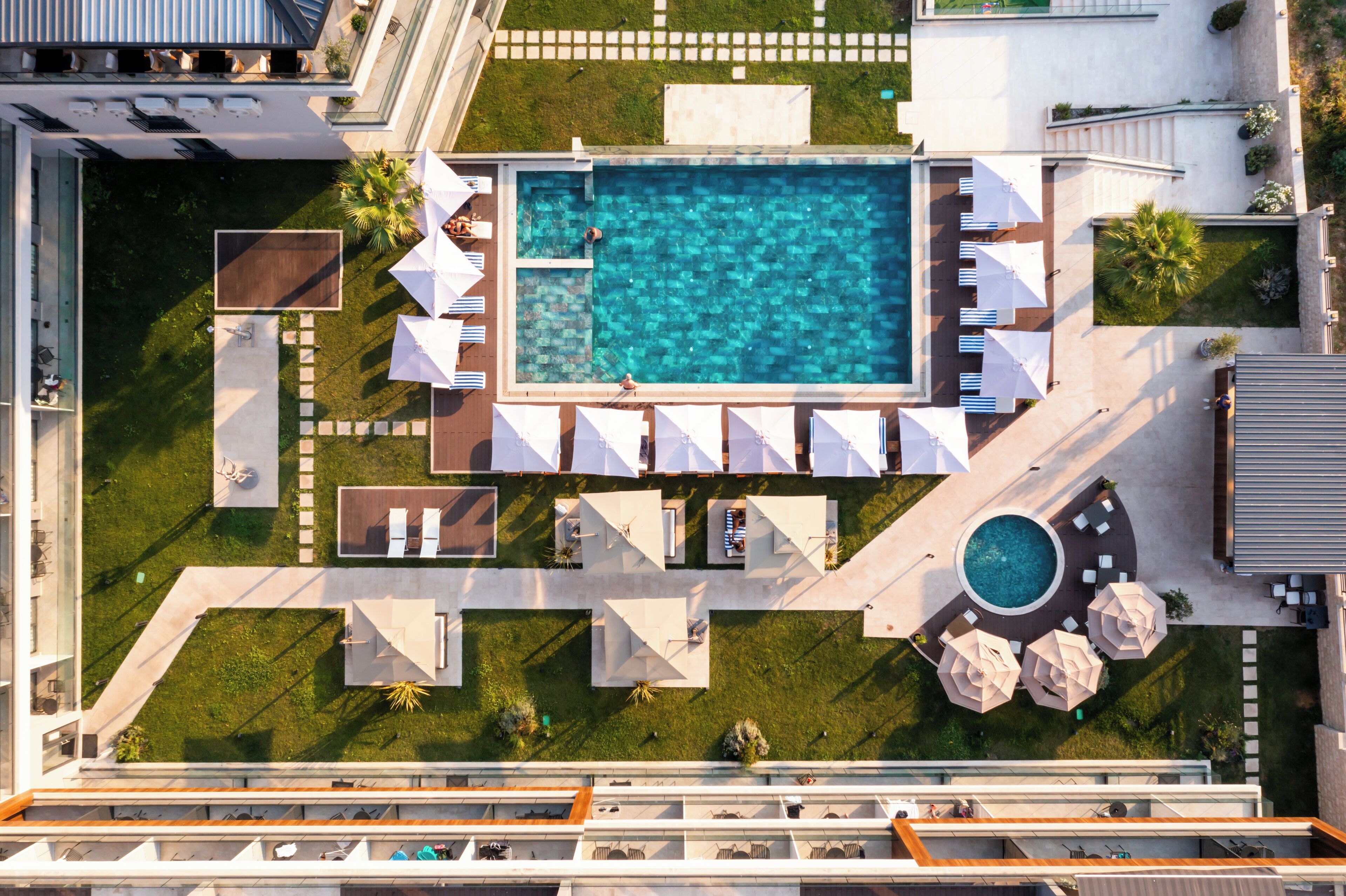 Eine Luftaufnahme eines Hotels mit einem zentralen Schwimmbecken, mehreren Liegebereichen mit Sonnenschirmen und einem kleinen, runden Pool.